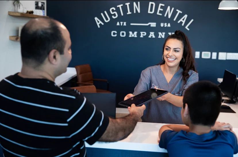 A smiling woman with long brown hair sits at a reception desk, holding a phone to her ear with her left hand and gesturing toward a computer screen with her right hand.
