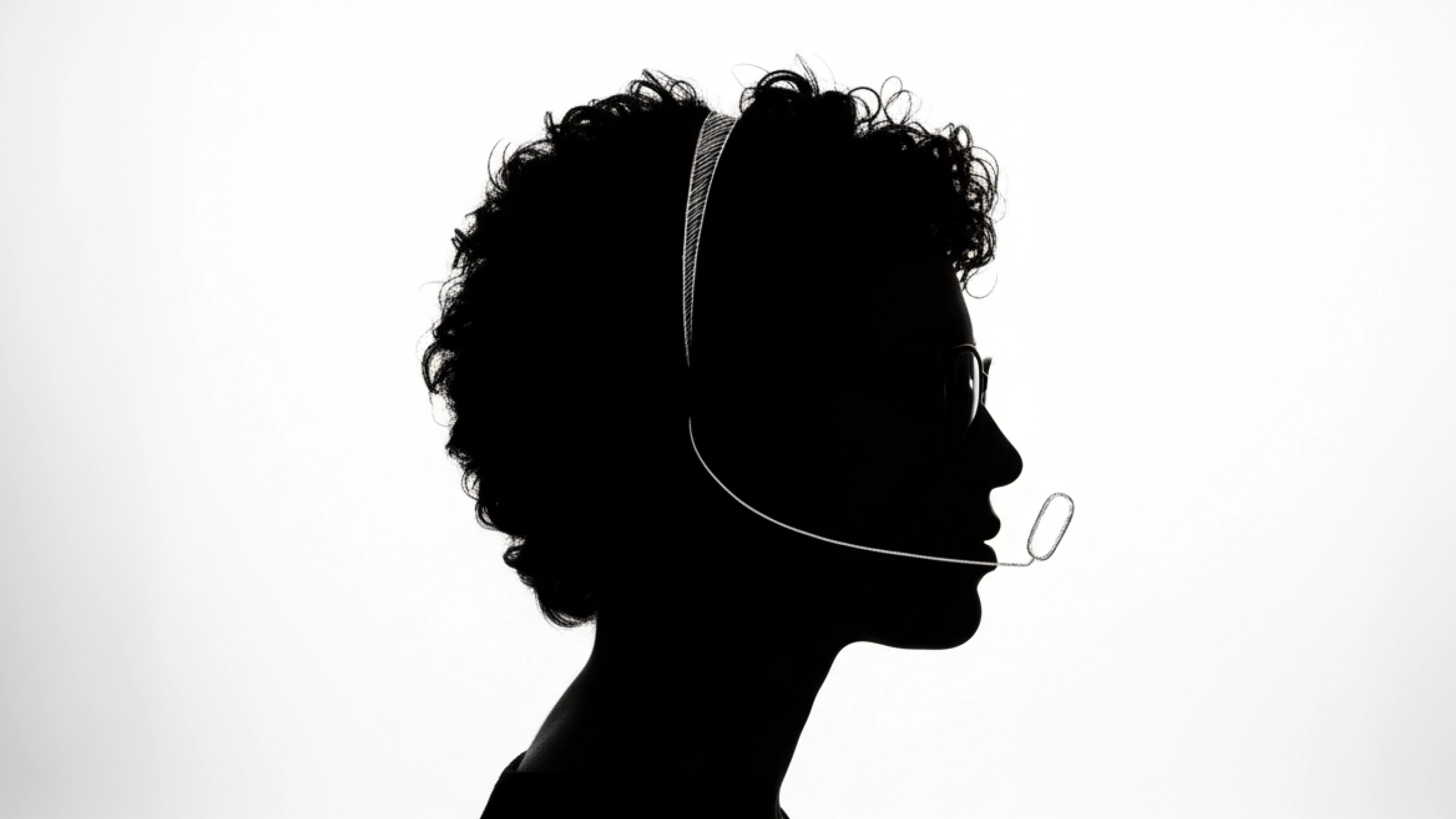 Silhouette of a person wearing glasses and a headset with a microphone against a bright background.