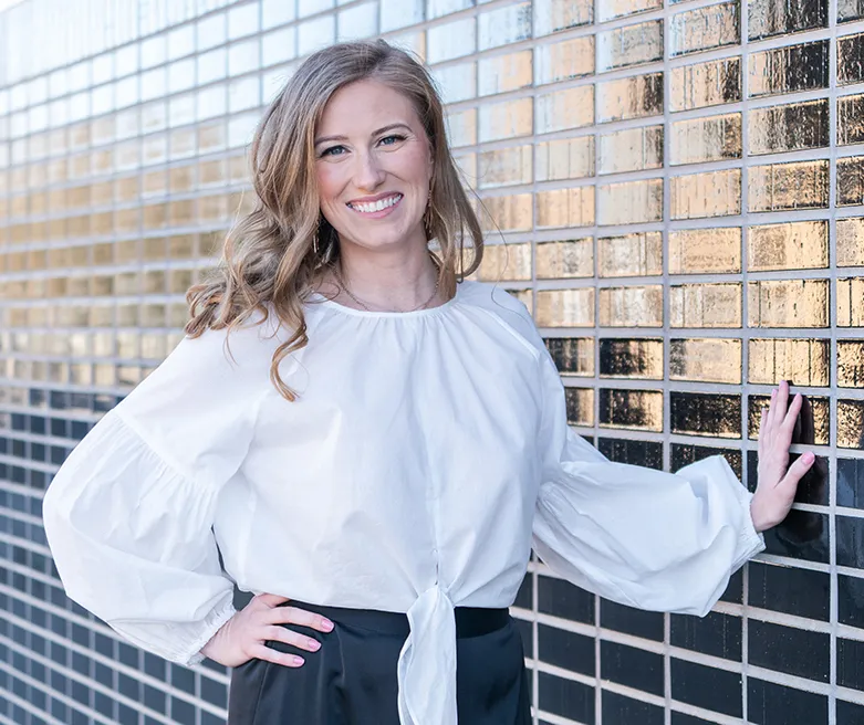 Blonde woman in a white long sleeve shirt smiling next to a reflective navy tile exterior wall
