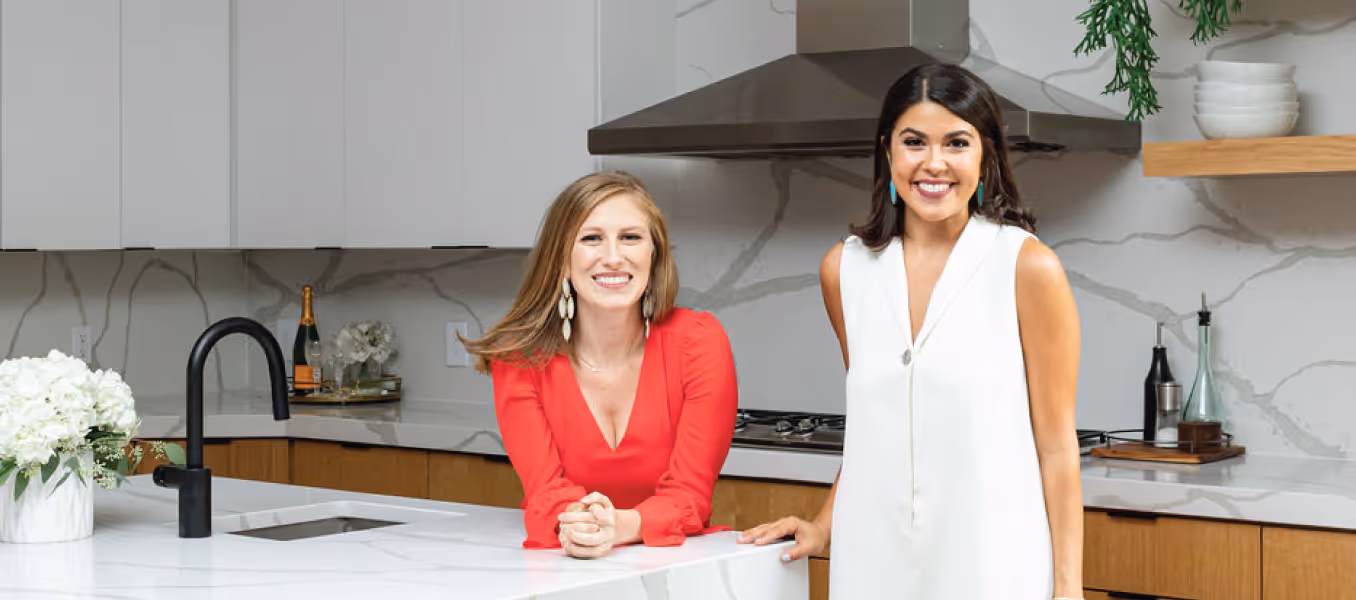 Two women posing together and smiling in a kitchen with a marble backsplash 