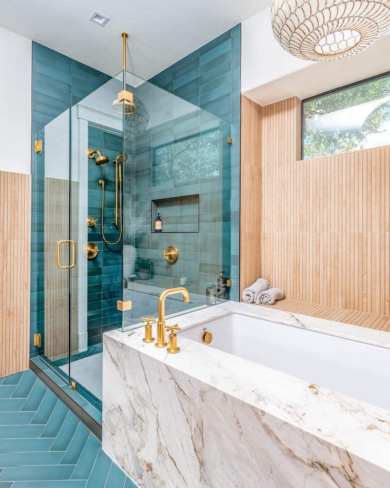 Bathroom with teal tiled shower with glass doors and a large tub with marble sides and a wood wall and chandelier 