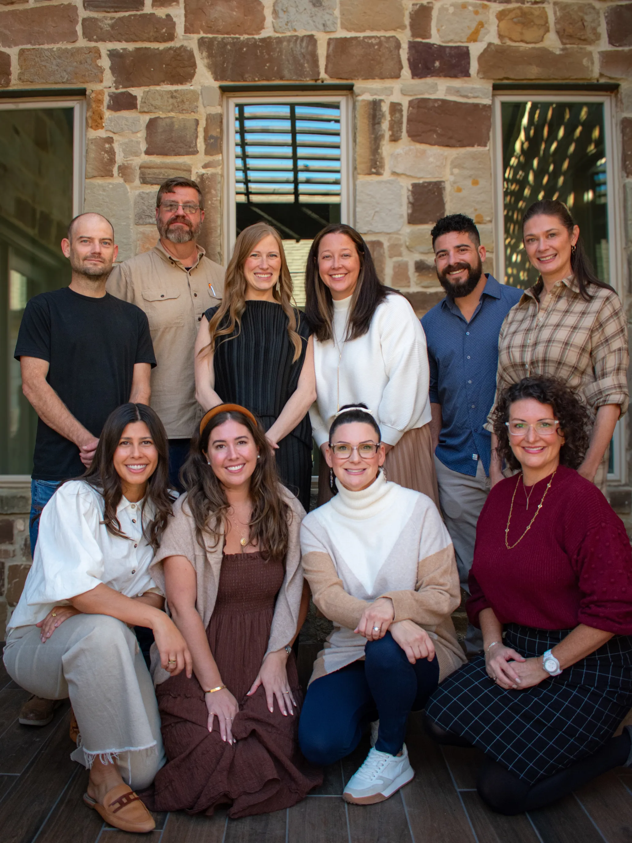 Group of ten adults, five standing and five kneeling, smiling in front of a stone wall with windows.