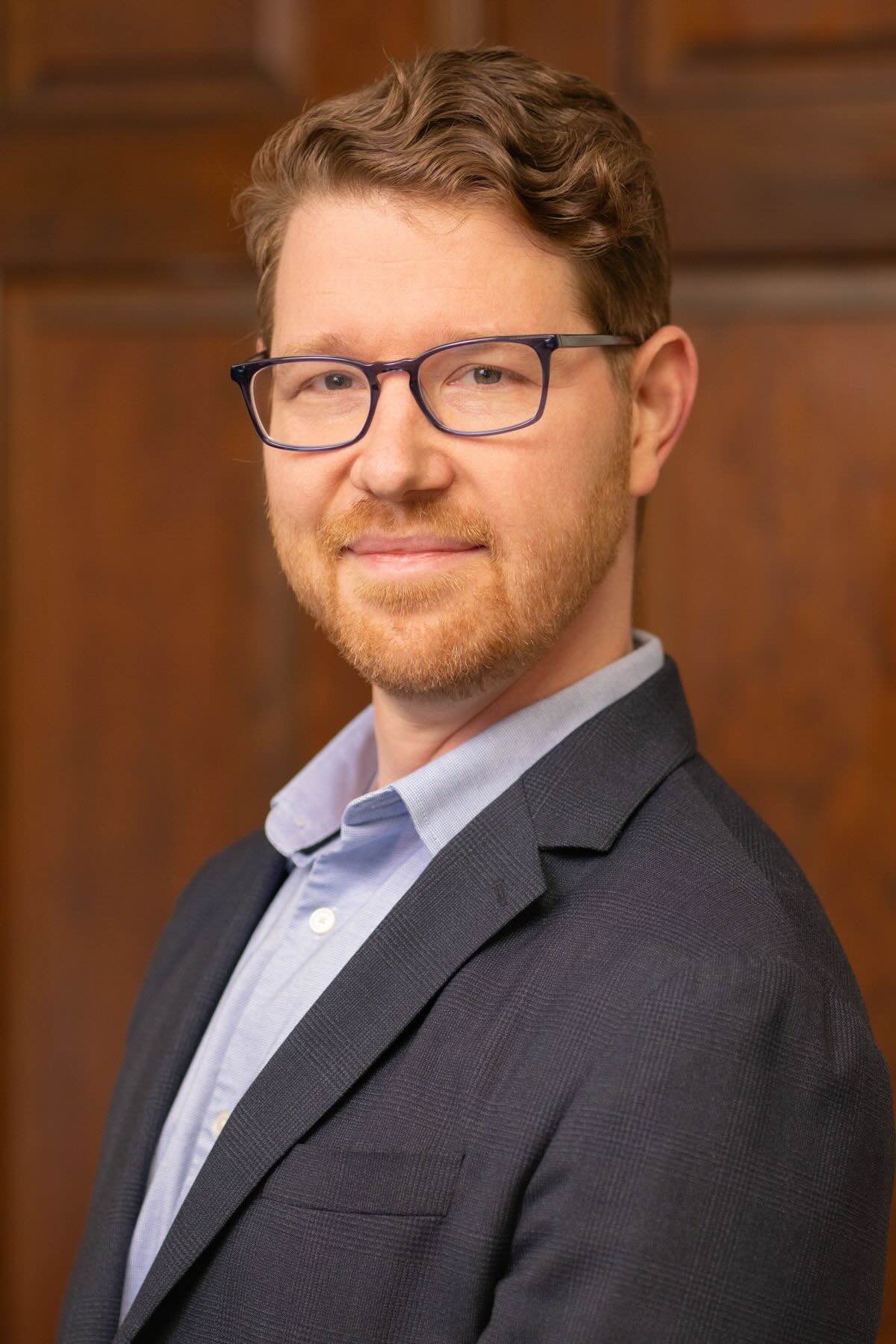 Portrait of a male audiologist with brown hair, glasses, and a beard wearing a white lab coat, blue dress shirt, and navy tie.