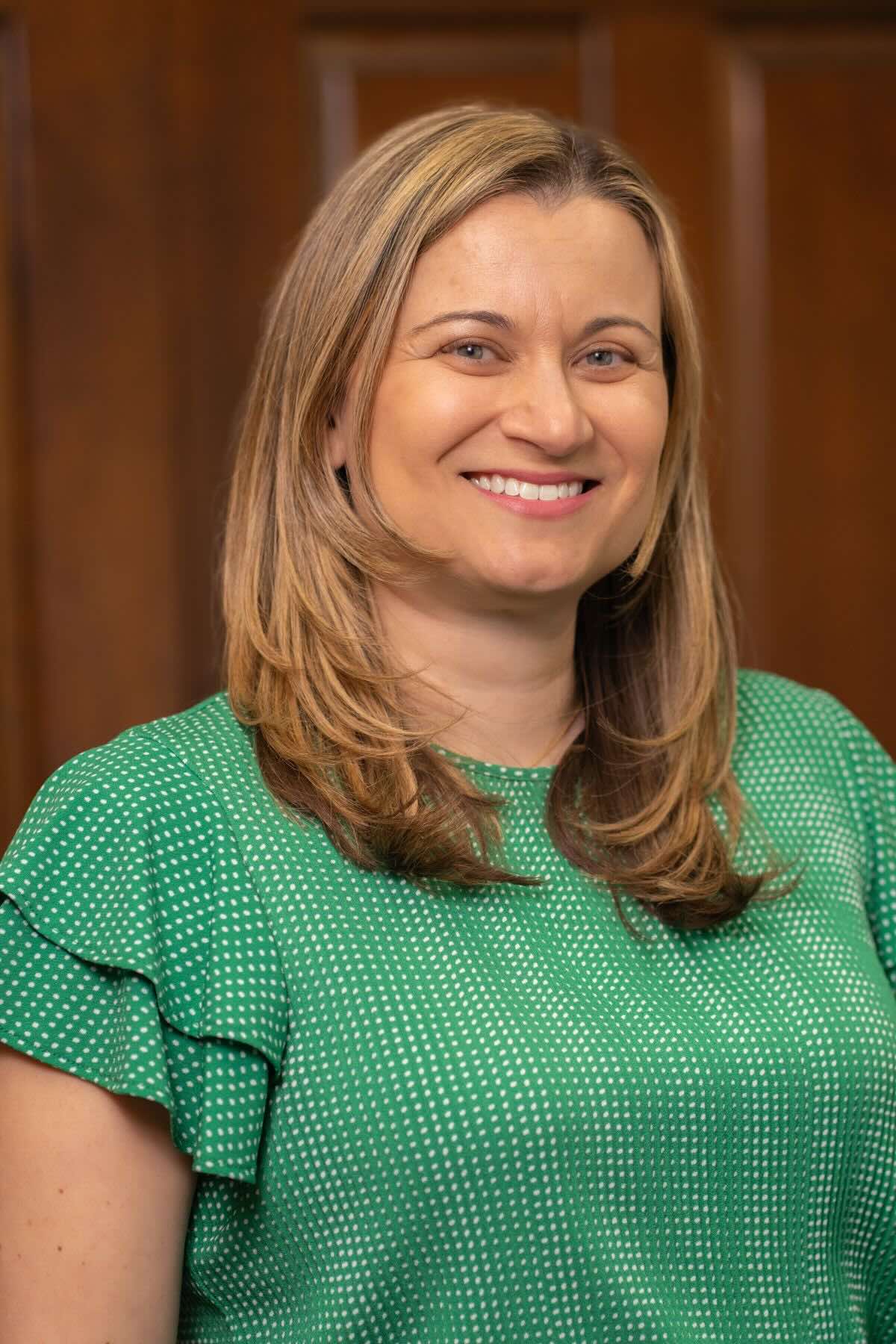 Close-up of a smiling female audiologist with light blue eyes and long brown hair wearing a mustard yellow top.