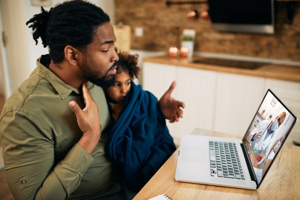 a dad and sick child sitting in front of their computer screen for a telehealth appointment