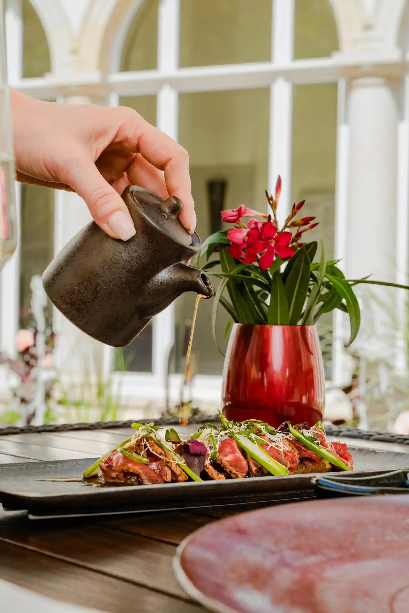 Hand gießt Sauce aus Ton-Kännchen über Rinder-Tataki mit Oleander – Villa Kennedy by Henssler