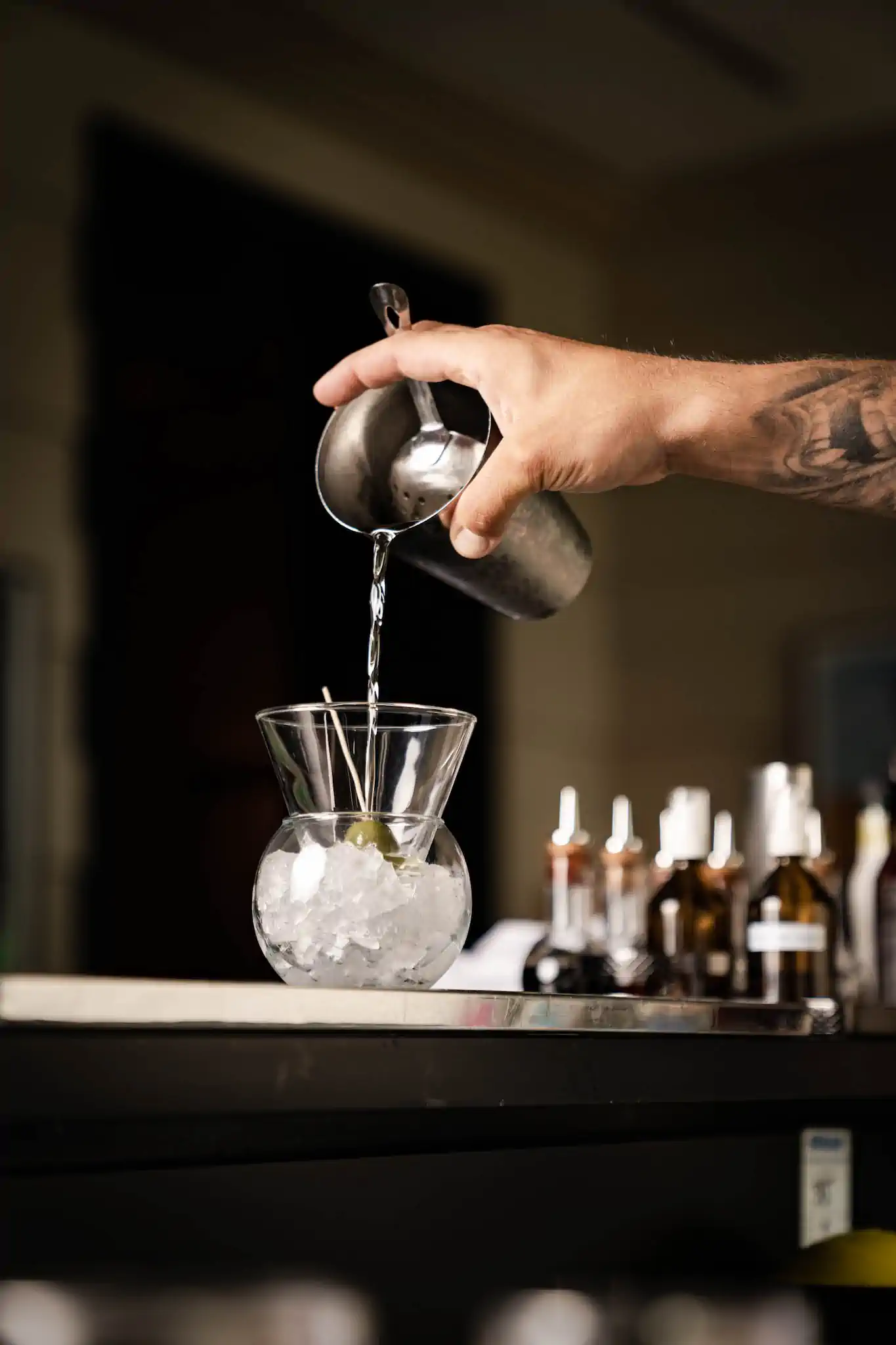 Tätowierter Unterarm gießt klaren Drink aus Shaker in Martini-Glas mit Olive – Hotel de Rome Berlin Bar
