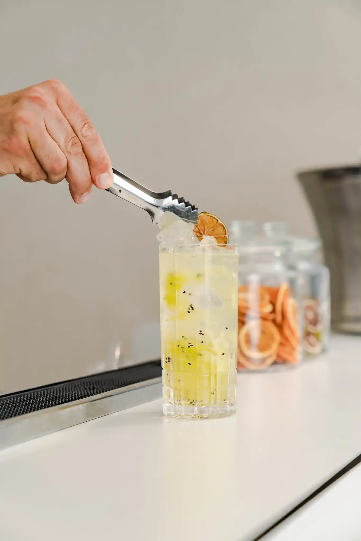 Barkeeper-Zange platziert getrocknete Limette auf Highball mit Kiwi-Slush – Hotel de Rome Berlin Bar