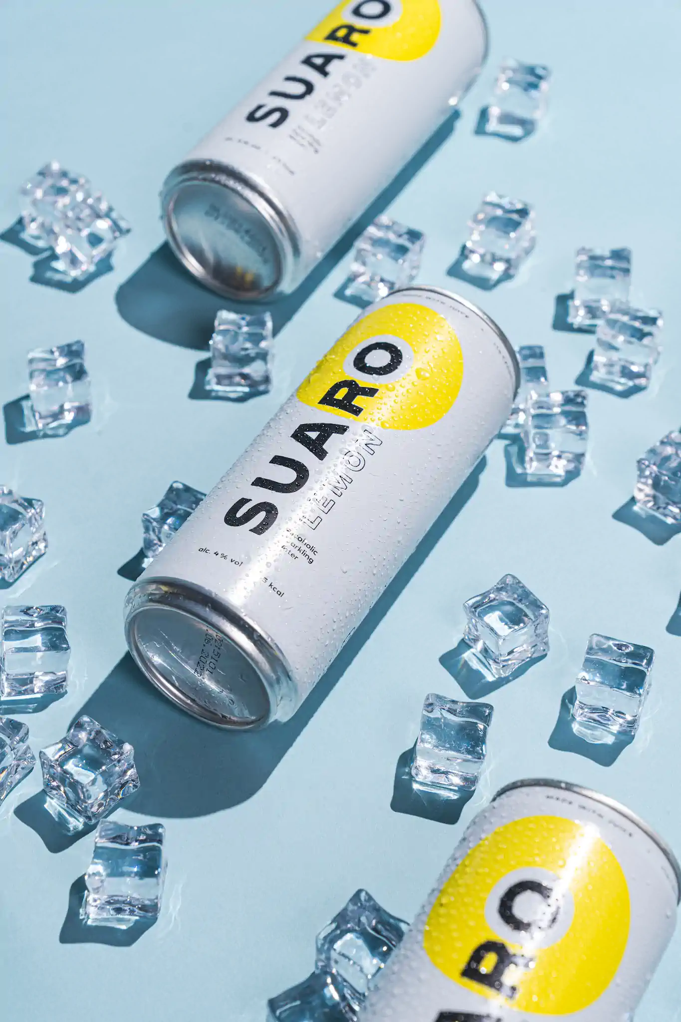 Zwei SUARO Lemon Dosen auf hellblauem Untergrund mit gestreuten Eiswürfeln – SUARO Getränkeshooting