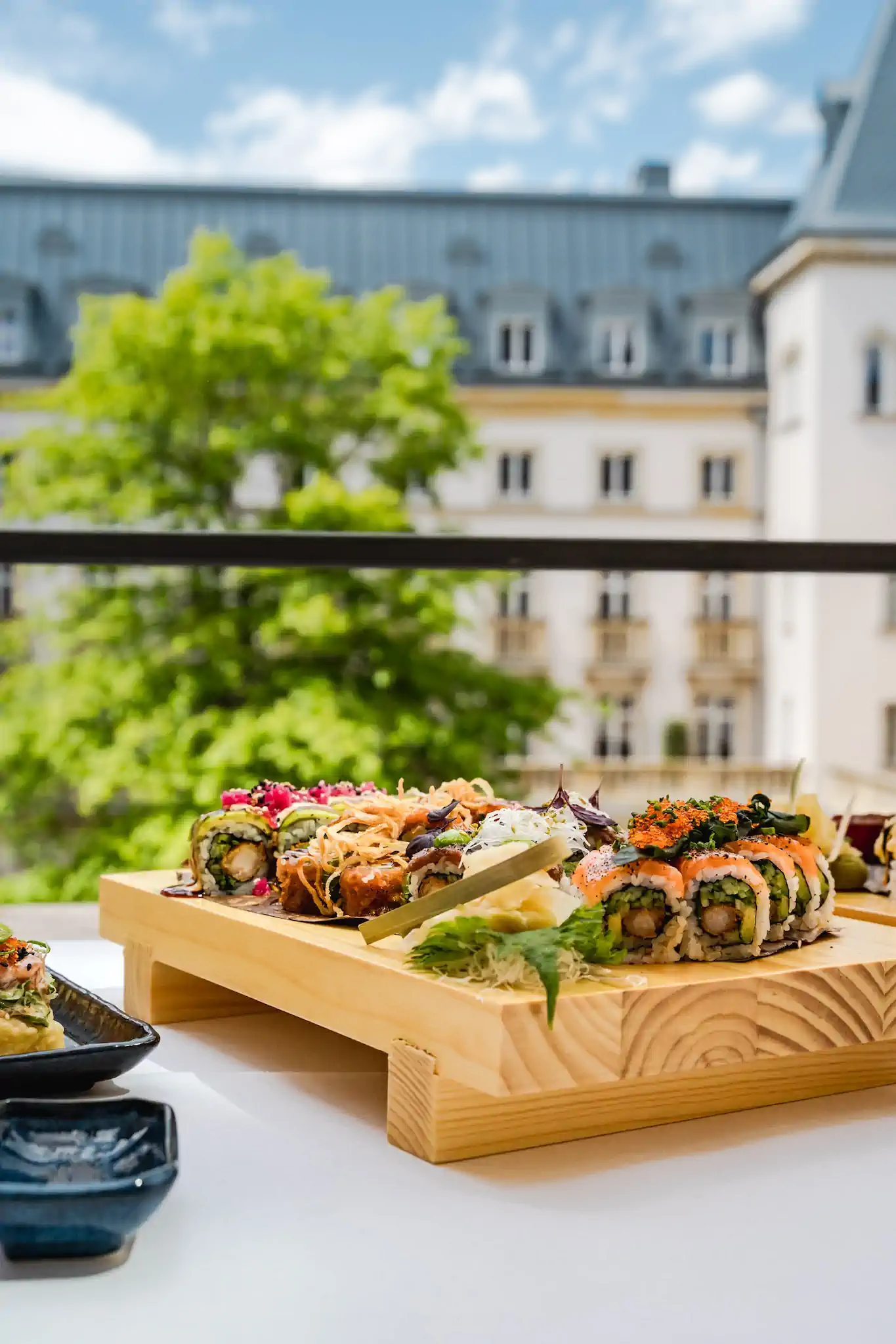 Sushi-Platte auf Holzboard vor barocker Villa-Kennedy-Fassade im Sommer – Villa Kennedy by Henssler