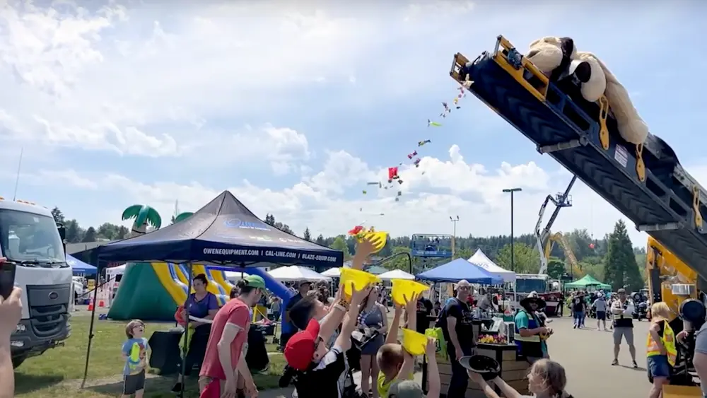 Children hold plastic hardhats over their heads to capture falling candy from a piece of heavy machinery.