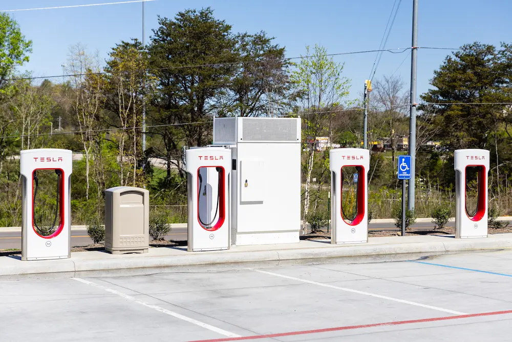 Four red and white Tesla branded EV chargers in a parking lot with trees and a road in the distance on a clear day.