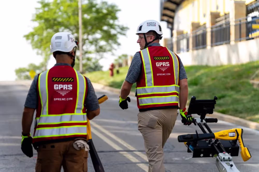 Two GPRS Project Managers walking uphill on a road carrying electromagnetic locators and pulling a ground penetrating radar scanning cart.