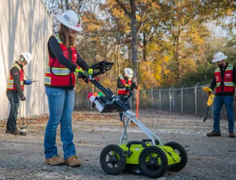 GPRS Project Managers, wearing work clothes, red safety vests and PPE, deploy various subsurface investigation and mapping technologies between a building and a fence. The worker in the foreground is using a walk-behind GPR unit, two workers in the mid-field of the photo are deploying an elephant’s foot amplifier for acoustic leak detection and an electromagnetic locator, while the worker in the background utilizes a GNSS GPS device.