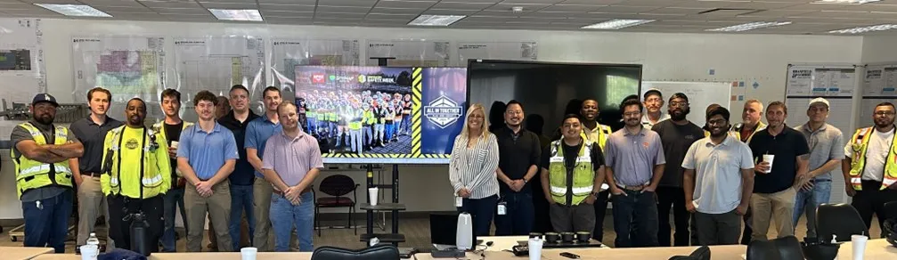 A group of people, some wearing yellow safety vests, standing side by side around a television with the Construction Safety Week promotional images shown