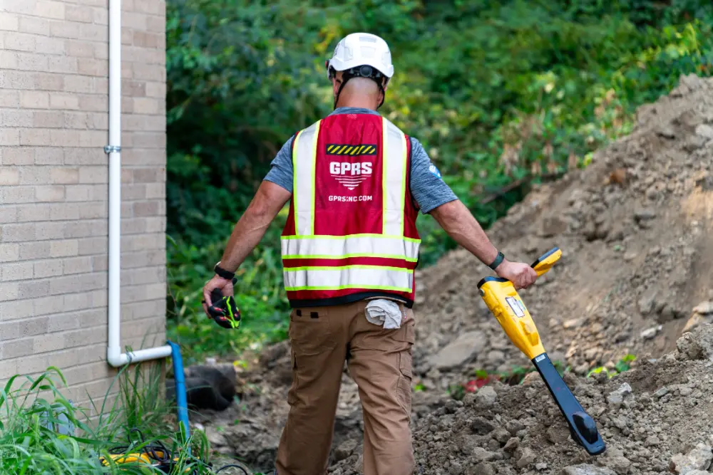 A GPRS Project Manager running an electromagnetic locator over a pile of dirt next to a building.