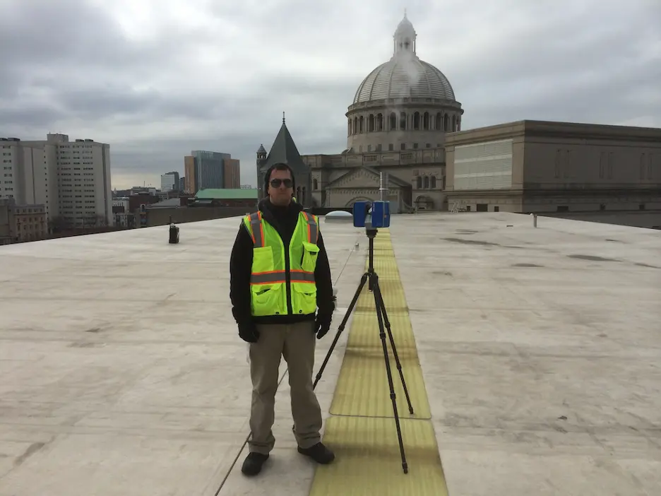 Kurt Yeghian pictured on a rooftop wearing a high-visibility safety vest and utilizing a 3D laser scanner on a tripod.