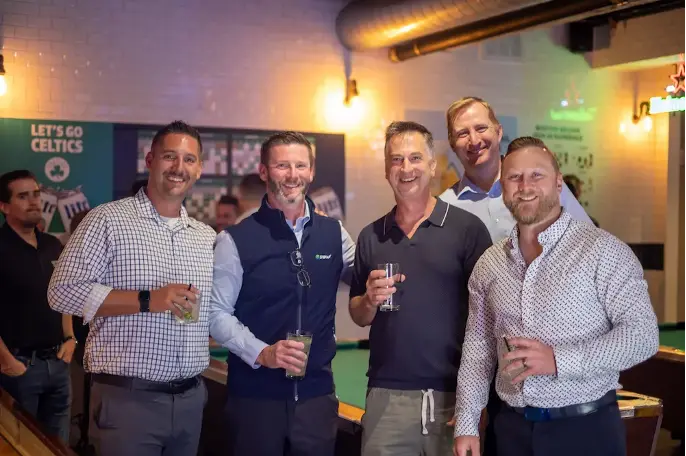 From left to right: Dave Mulcahey, Vice President of Sales, Steve Mumm, Chief Commercial Offer, Yeghian, Dustin Snavely, Vice President of National Sales, and Adam Yunker Vice President of Commercial Operations standing together holding drinks. A 'Let's Go Celtics' sign is visible in the background along with various decorations.