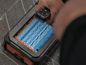 A handheld ground penetrating radar device that shows wavy lines in the shape of hyperbolas. The ground penetrating radar is held in a human hand and placed on top of a concrete slab with white lines painted on it, representing concrete rebar. 