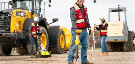 A GPRS Project Manager in the foreground utilizing an electromagnetic locator. In the background, there are two other Project Managers, one using ground penetrating radar and the other is using the electromagnetic transmitter. They are all standing in front of two bulldozers on a dirt job site.