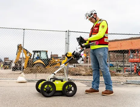 A GPRS Project Manager utilizing ground penetrating radar on a job site in front of a fence. Behind the fence is a job site with an orange building and excavators.