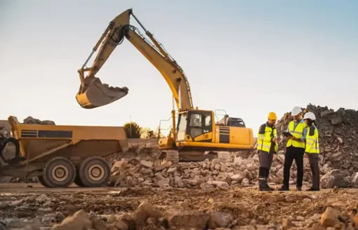 An excavator on a dirt job site with three construction workers standing to the left of it wearing helmets and bright yellow vests while looking at an iPad together.
