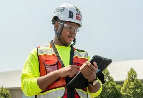A GPRS Project Manager looking at an iPad they are holding while wearing proper PPE including a red vest, protective glasses and a helmet.