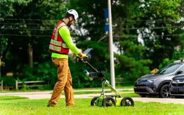 A GPRS Project Manager pushing ground penetrating radar across a grassy field with trees and cars in the background.