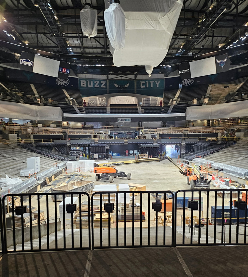 A photo of the inside of the Spectrum Center that is under construction. Plastic wrap is covering the jumbotron and other LED displays. Construction equipment is where the basketball court would be that is now a concrete floor. The Charlotte Hornets logo is seen at the top of the back wall with the words “Buzz” and “City” on the left and right of it respectfully.