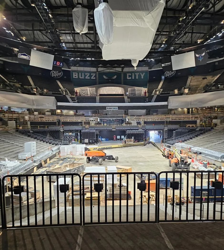 A photo of the inside of the Spectrum Center that is under construction. Plastic wrap is covering the jumbotron and other LED displays. Construction equipment is where the basketball court would be that is now a concrete floor. The Charlotte Hornets logo is seen at the top of the back wall with the words “Buzz” and “City” on the left and right of it respectfully.