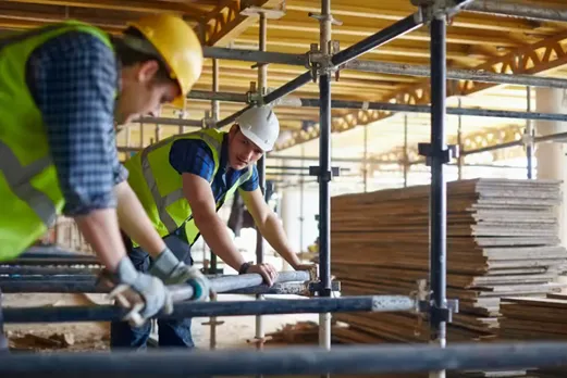 Two construction workers wearing vests and helmets while holding a pipe on a job site surrounded by many rows of pipes and construction materials.