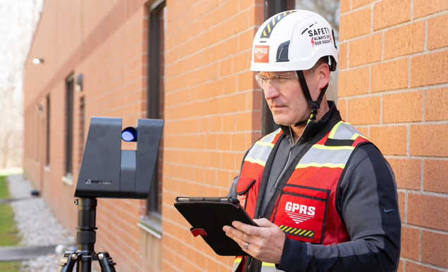 A man wearing a helmet and red vest holding an iPad while standing behind a Matterport Pro3 and in front of a brick wall.
