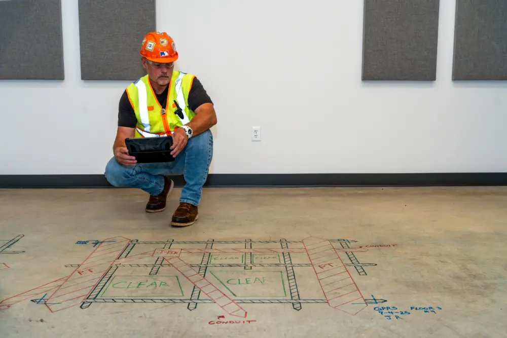 A construction worker holding a tablet kneels in front of a concrete scanning mark-out on a slab.