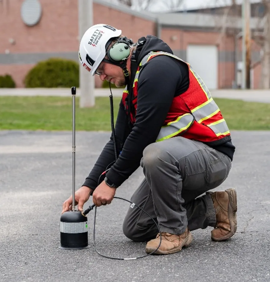 A GPRS Project Manager wearing a white helmet, green noise-cancelling headphones, a black hoodie, grey pants, a red vest, and brown shoes setting up an acoustic leak detector on asphalt. 