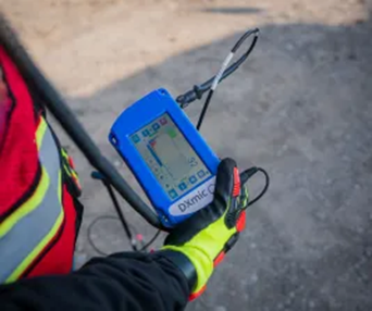 A person wearing a bright yellow, red and black glove and a bright yellow, red and grey safety vest holding a blue leak noise correlator with a grey screen.