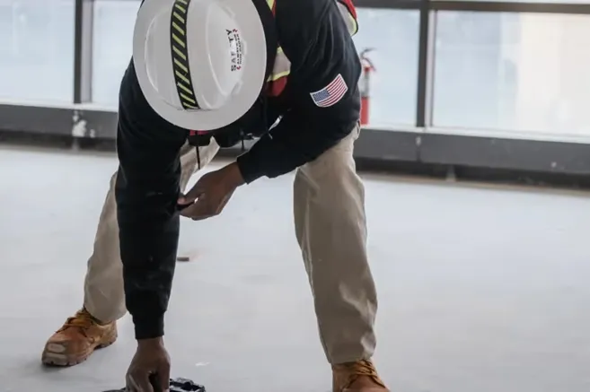 A man wearing a white safety helmet, red vest, black long sleeve shirt, and khaki pants scanning concrete with a handheld GPR concrete scanner. 