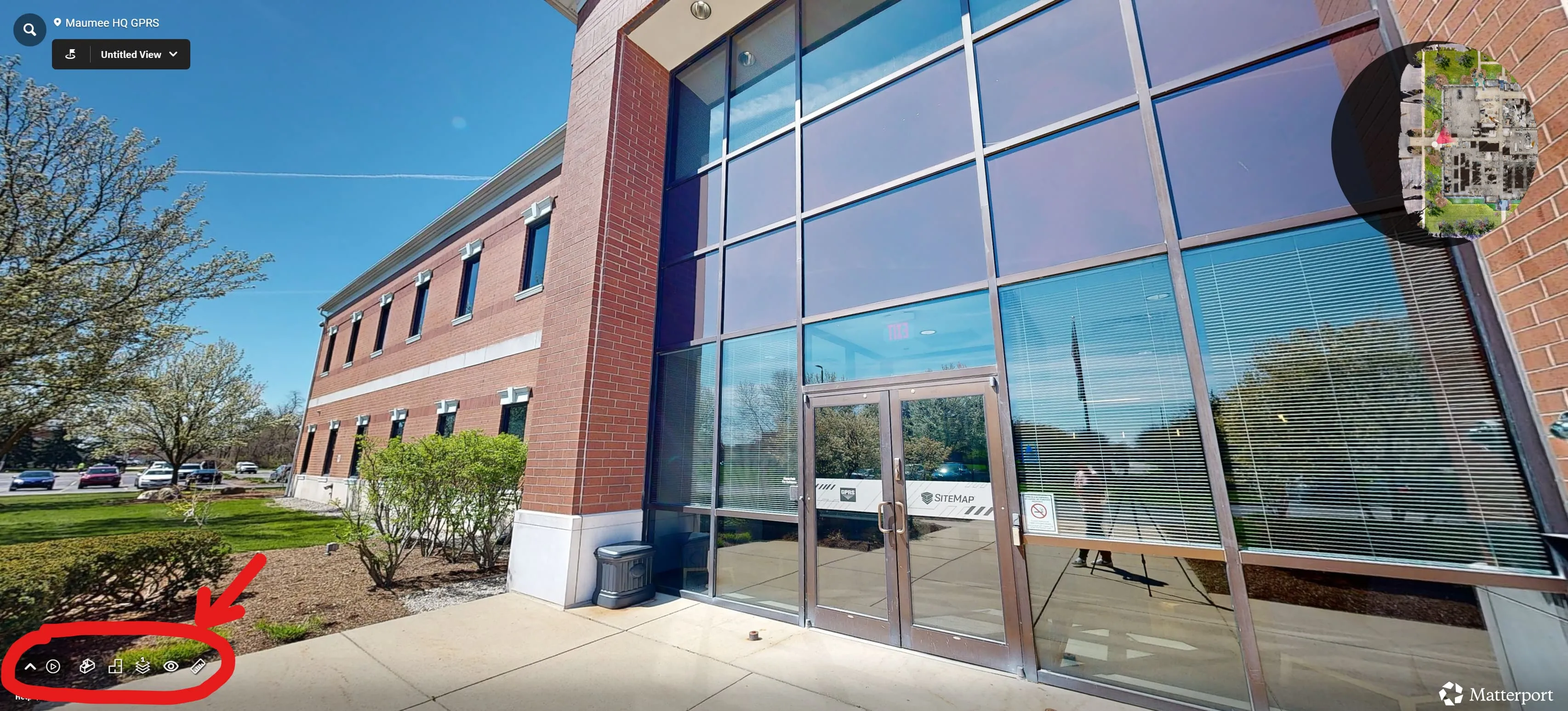 A virtual tour from outside a multi-story brick building with an aerial view of it in a circle in the top right corner. Tools in the bottom left corner are shown within a red circle with a red arrow pointing at it. 
