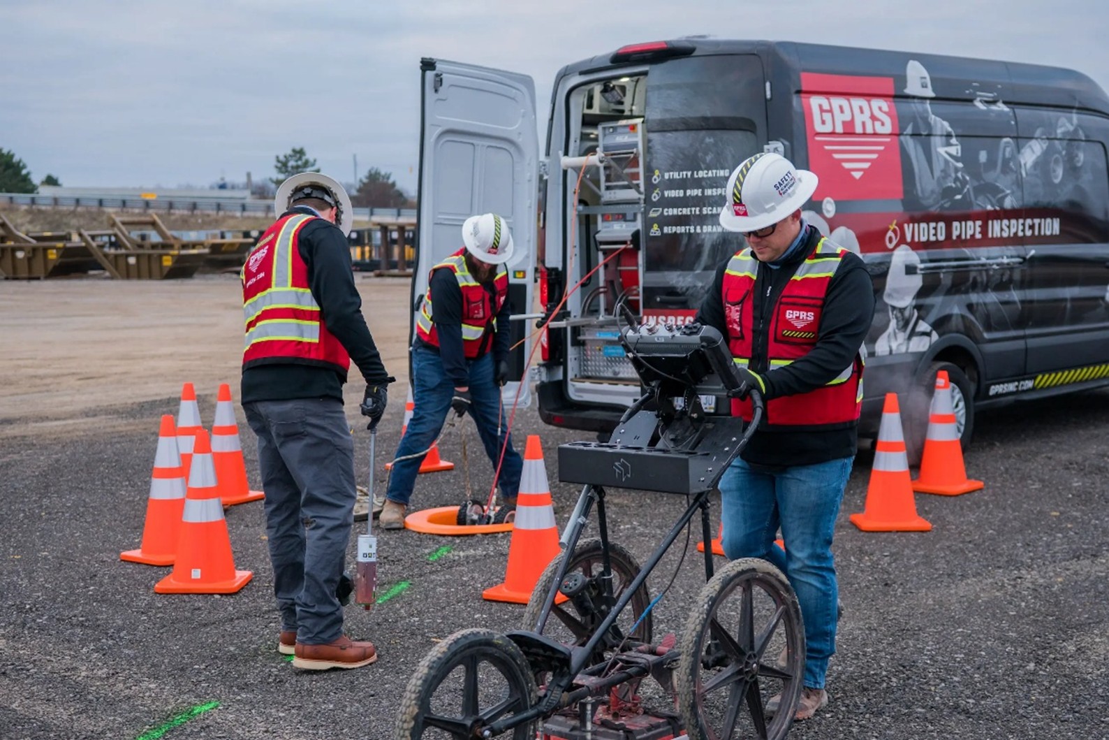 Three GPRS workers wearing white hard hats, red and yellow vests, black sweater long sleeved shirts, and jeans marking out utilities and lowering a VPI crawler into a manhole surrounded by eight orange cones by a grey GPRS truck.