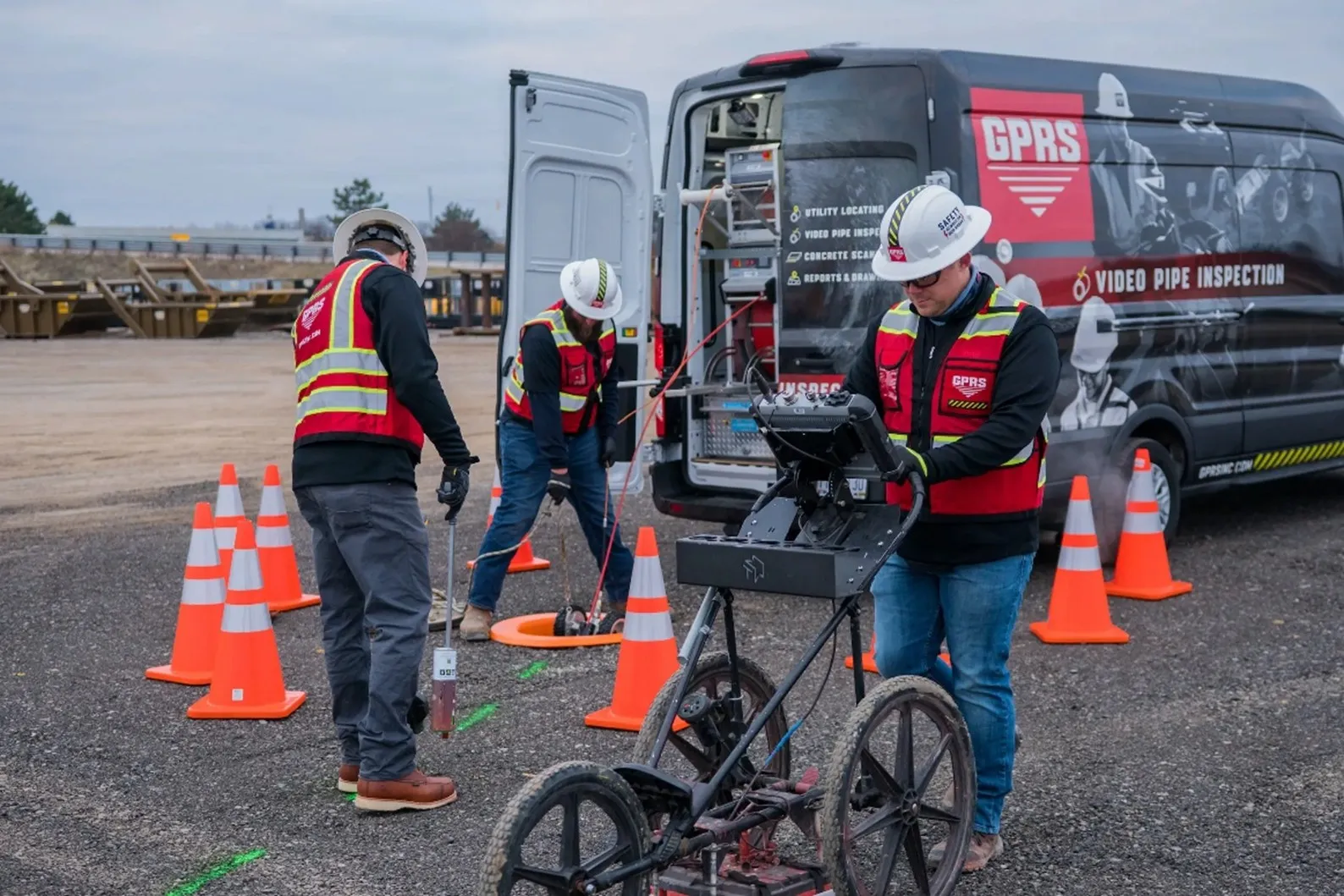 Three GPRS workers wearing white hard hats, red and yellow vests, black sweater long sleeved shirts, and jeans marking out utilities and lowering a VPI crawler into a manhole surrounded by eight orange cones by a grey GPRS truck.