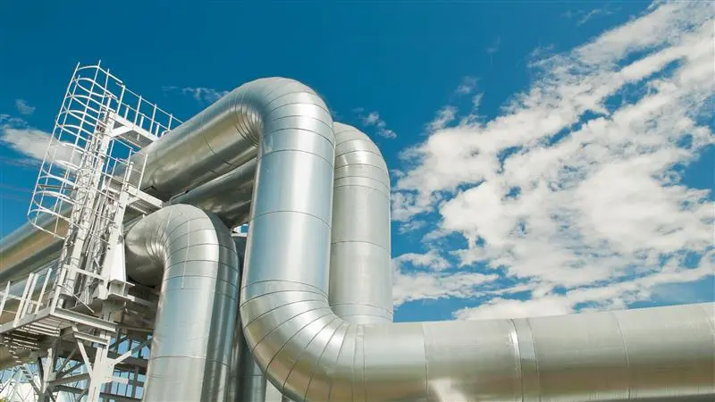 New brightly shining sliver natural gas pipelines bend in elbow joints above the ground, with a white-painted access ladder & platform, framed against a blue sky with light clouds.