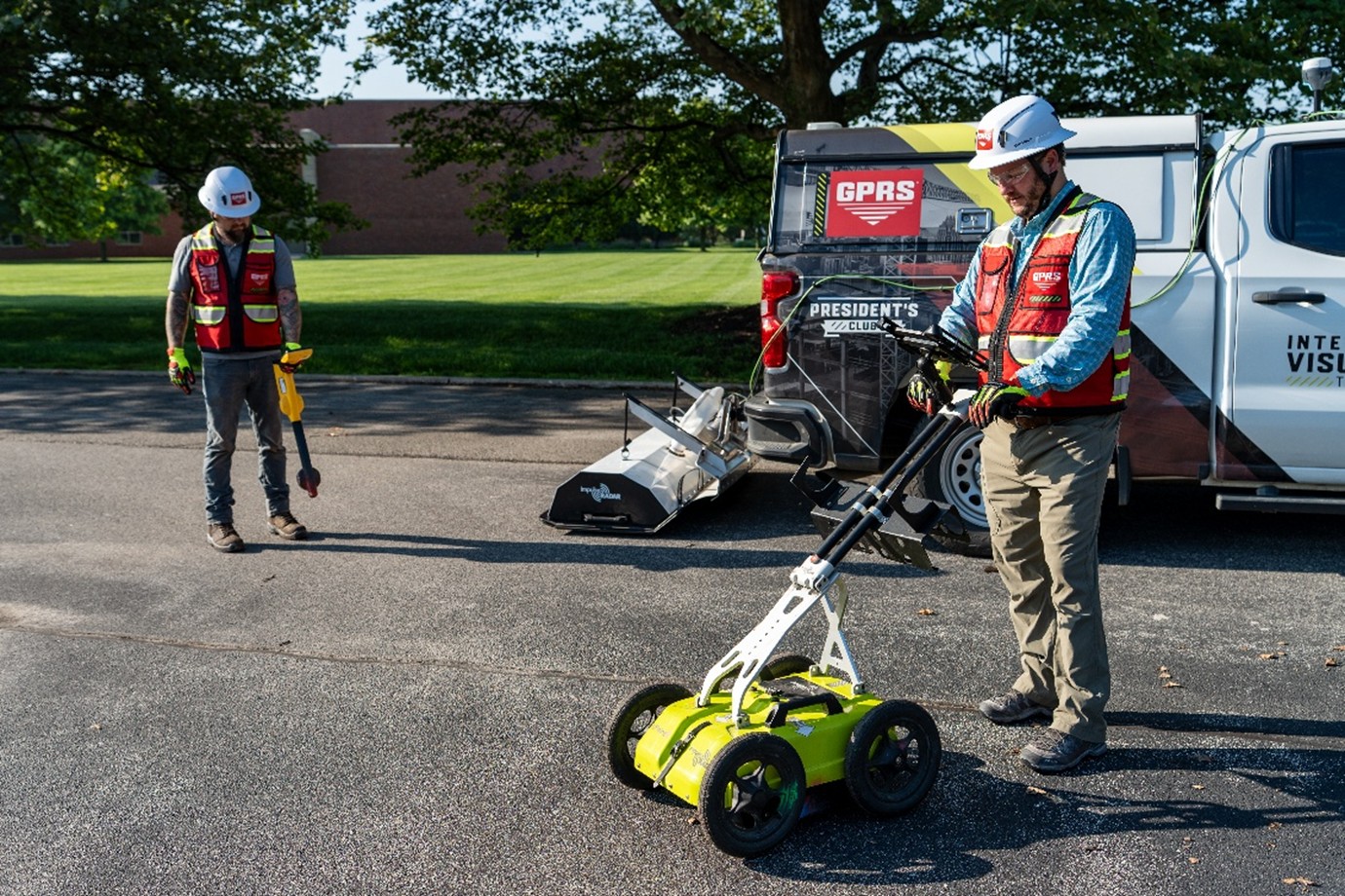 Two GPRS Project Managers wearing a white helmet and a red vest. One is standing to the right of the screen using the GPR push cart and the other is standing to the left using an electromagnetic locator. In between them is a GPRS truck with a GPR array attached to the back of it.