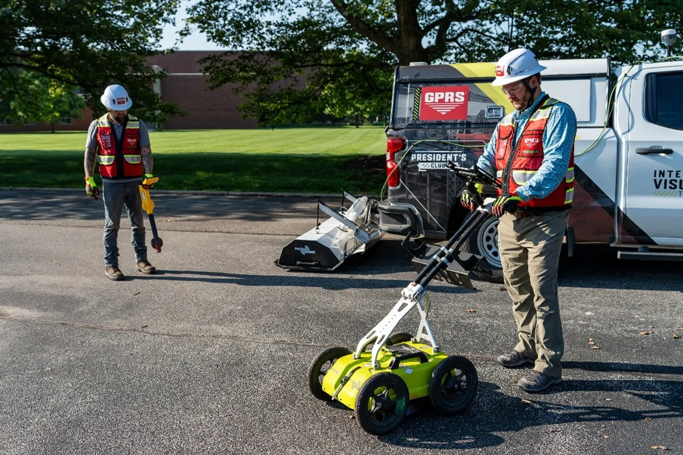 Two GPRS Project Managers wearing a white helmet and a red vest. One is standing to the right of the screen using the GPR push cart and the other is standing to the left using an electromagnetic locator. In between them is a GPRS truck with a GPR array attached to the back of it. 