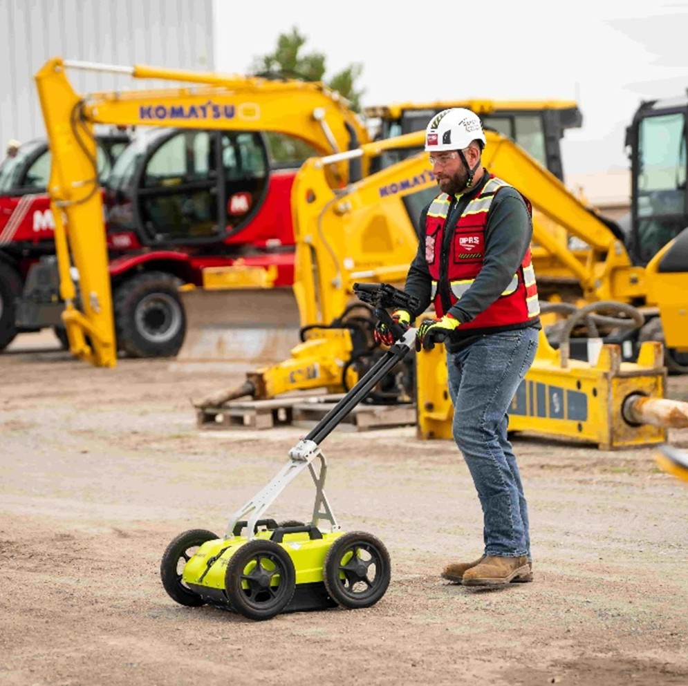 A GPRS Project Manager using a bright yellow, silver, and black GPR pushcart in front of construction equipment on a dirt job site.
