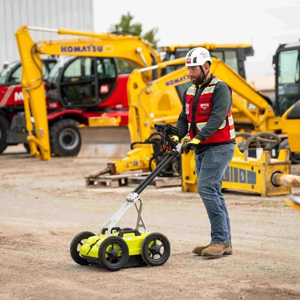 A GPRS Project Manager using a bright yellow, silver, and black GPR pushcart in front of construction equipment on a dirt job site.  