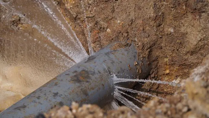 A gray underground pipe surrounded by dirt spraying out water from a crack in the pipe.