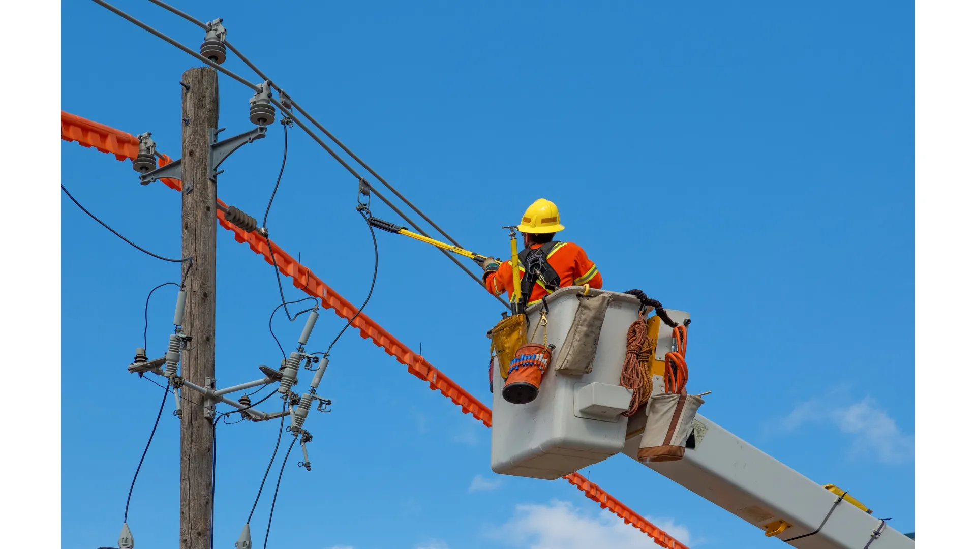 A construction worker wearing high-visibility clothing and a hard hat on a lift assessing a power line. 