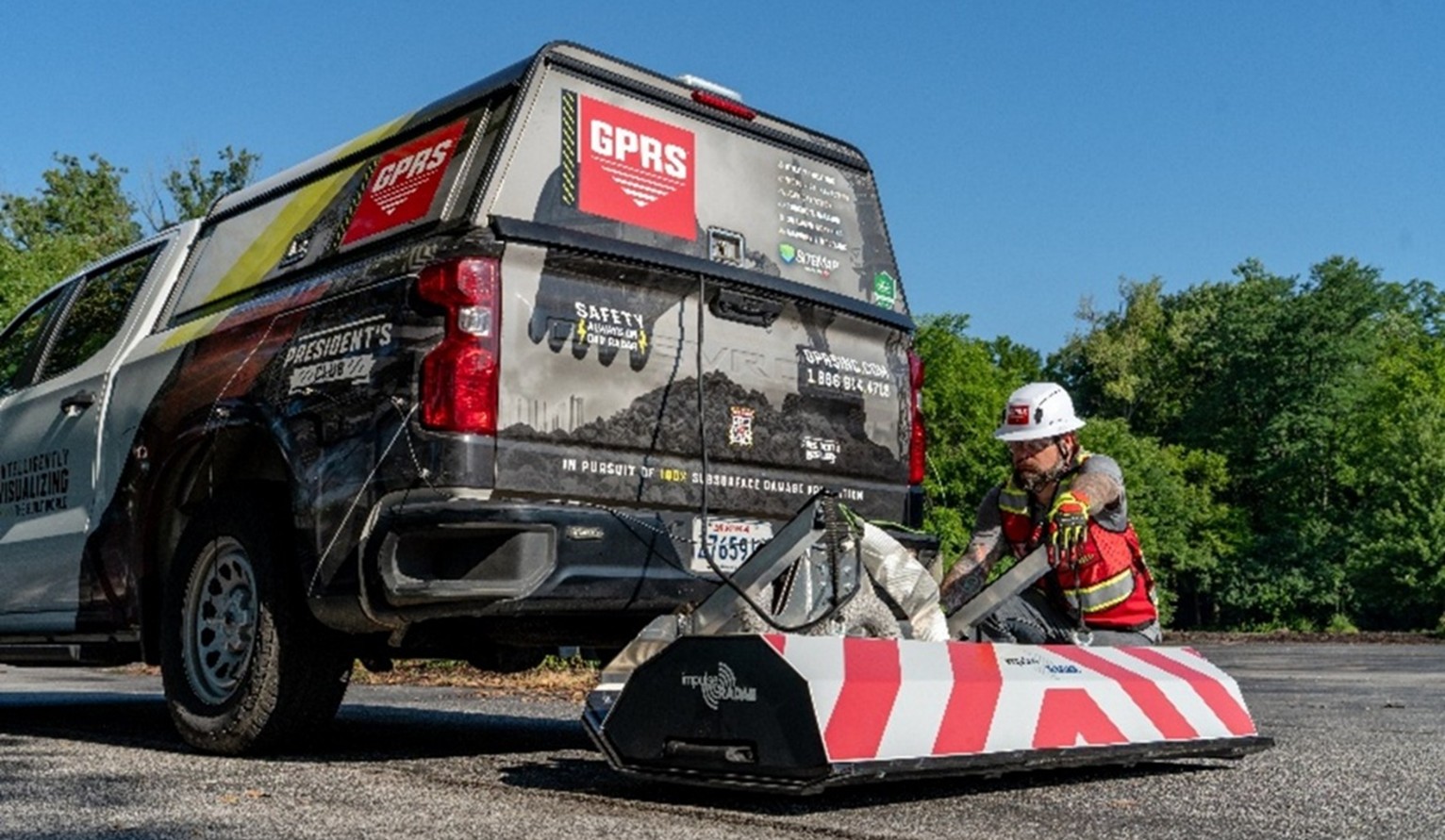 A truck with GPRS branding with a High-Speed 3D GPR array with white and red stripes being attached by a SIM-certified GPRS Project Manager wearing the proper PPE, like a hard hat and red vest.