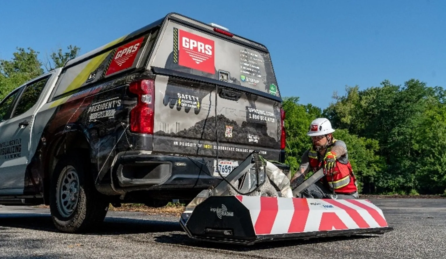 A truck with GPRS branding with a High-Speed 3D GPR array with white and red stripes being attached by a SIM-certified GPRS Project Manager wearing the proper PPE, like a hard hat and red vest.