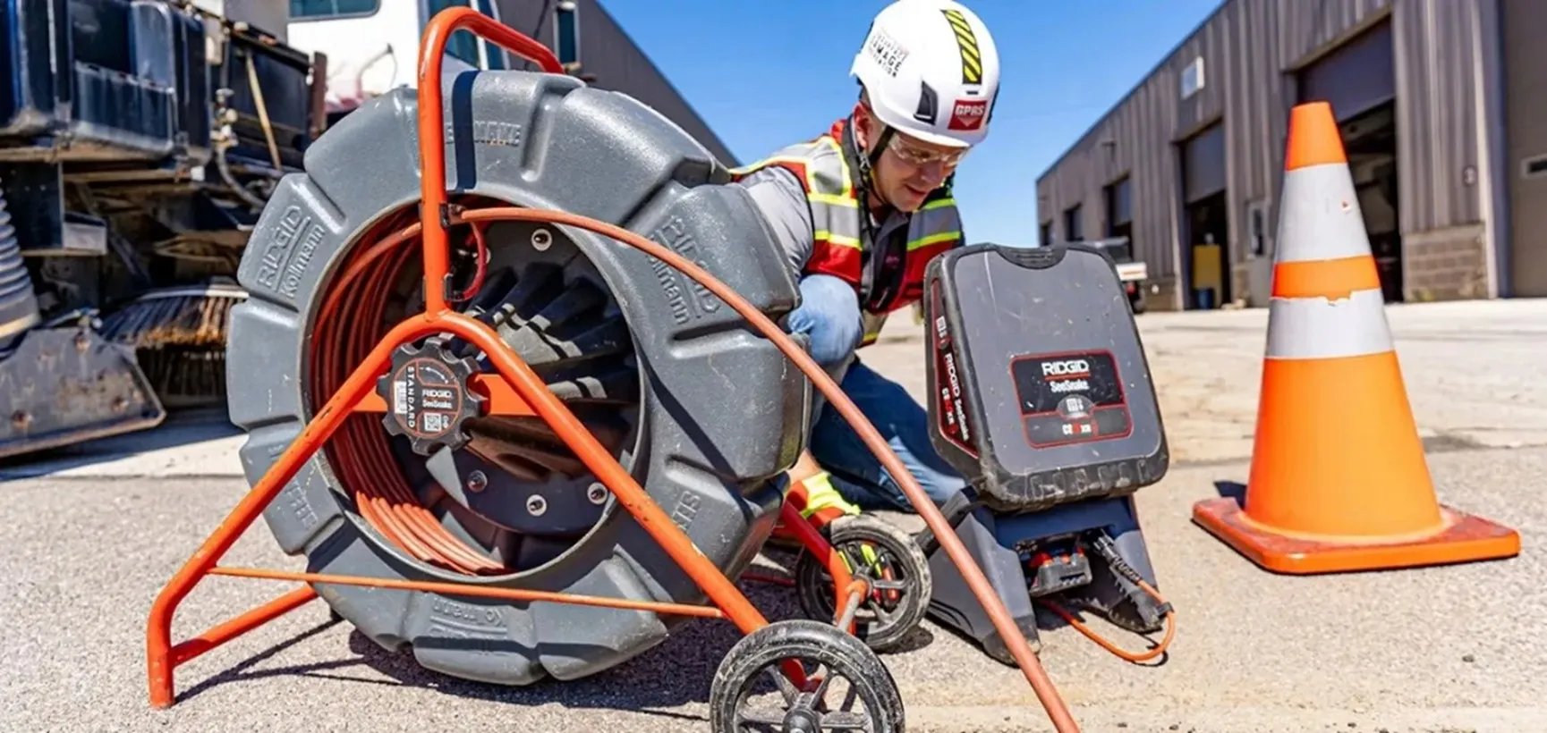 A GPRS Project Manager wearing a helmet & red vest while utilizing a push camera by lowering it into a manhole. There is also an orange cone to the right of the Project Manager. 