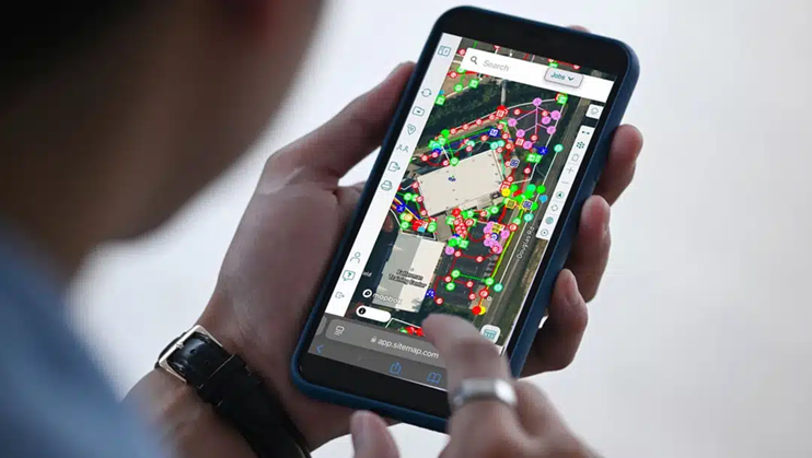 A smartphone being held by the left hand wearing a watch while the right hand that has a ring on the pointer finger is pointing at the screen, which shows a satellite image, toolbars on the left & right side, and green, red, pink, orange, and blue lines representing underground utilities.